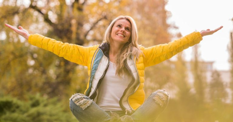 woman in yellow jacket and ripped jeans sitting cross-legged with her arms outstretched and looking up to the sky smiling